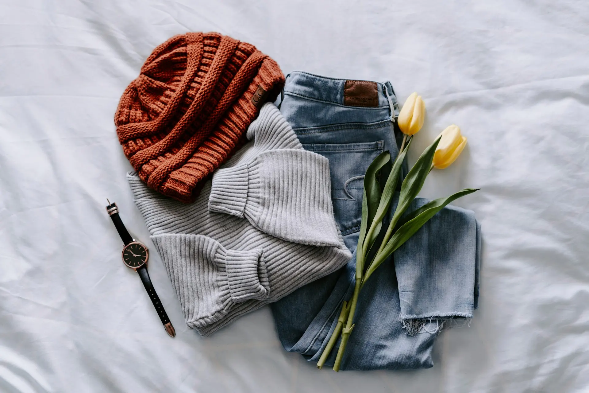 Pullover, Jeans, Mütze und Uhr auf einem weißen Bett ausgelegt